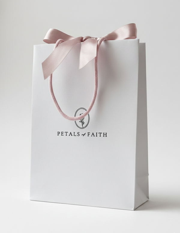 Photograph of white gift bag with dusty pink satin bow ribbon and pink cotton handles. Medium size with black embossed petals of faith logo