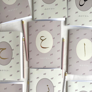 A flat-lay arrangement of multiple pastel pink and off white floral memo notebooks with gold Arabic calligraphy designs on the covers, interspersed with two pink and gold pencils on a light linen background.