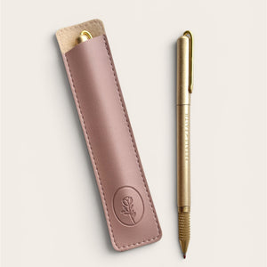 A gold-colored ballpoint pen with a gold clip and nib, displayed next to its matching dusty rose pink leather sleeve on a light beige background.