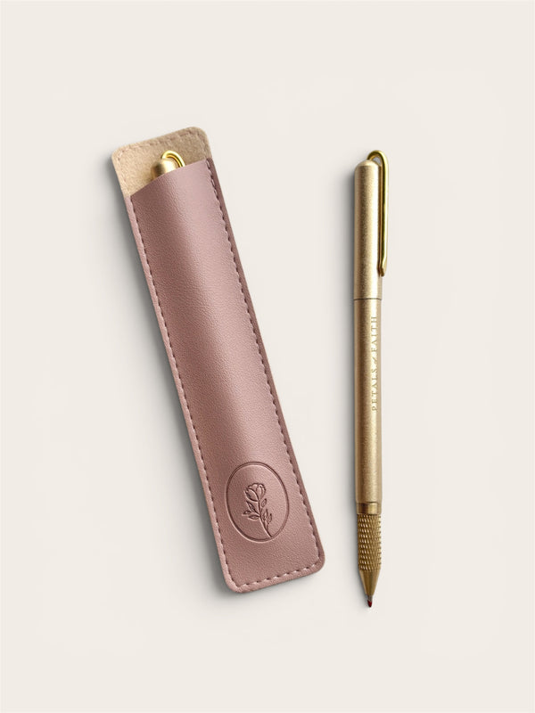 A gold-colored ballpoint pen with a gold clip and nib, displayed next to its matching dusty rose pink leather sleeve on a light beige background.
